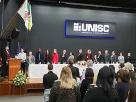 Auditório da Universidade de Santa Cruz do Sul. No centro da imagem, a mesa de abertura do evento composta por quinze, incluindo a presidenta do COEPEDE Jussara Muller de Assis. No fundo uma parede preta com o nome da Universidade. O palco, onde a mesa de