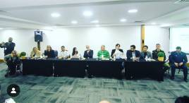 #DescriçãodaImagem Foto da mesa de abertura, fundo uma parede branca, luzes refletindo no teto, mesa longa, sob a mesa copos e garrafas de agua e sentados componentes do Ministério do Esporte, Secretaria Estadual de Esporte e Lazer, Secretaria Municipal