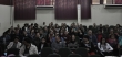 Na imagem, detalhe do auditório. As pessoas estão todas sentadas em poltronas verdes. 